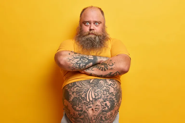 serious-surprised-blue-eyed-man-with-thick-beard-keeps-arms-folded-listens-wife-explanation-feels-jealous-has-big-tummy-overweight-because-wrong-nutrition-isolated-yellow-wall