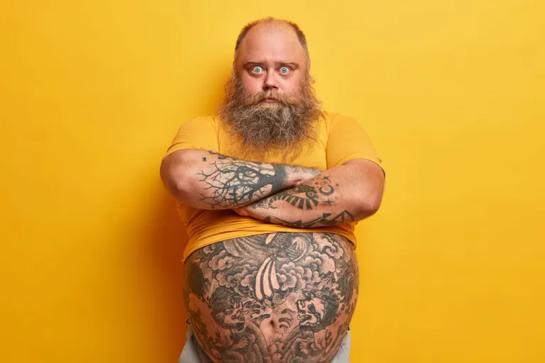 serious-surprised-blue-eyed-man-with-thick-beard-keeps-arms-folded-listens-wife-explanation-feels-jealous-has-big-tummy-overweight-because-wrong-nutrition-isolated-yellow-wall