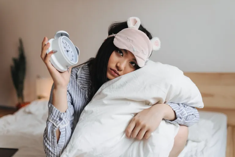 sad-girl-pajamas-sleep-mask-is-hugging-white-pillow-holding-alarm-clock