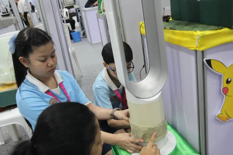 น้อง ๆ กำลังสาธิตการติด "แผ่นกรองฝุ่นจากผักตบชวา" บนพัดลมไร้ใบพัด